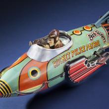 Side view of light green and orange rocket ship figure with character figure in cockpit. "Rocket Police Patrol" is painted on the side of the cockpit in yellow and black.