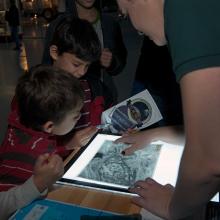 One staff member of the Museum and two younger-aged Museum visitors work together to solve a game. They are looking at an object on a white screen.