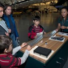 Four Museum visitors and one member of the Museum's staff work together to solve a game. Two visitors and the staff member are playing with a silver-colored twisted object.