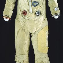 Front view of full-body spacesuit made of tan fabric. Suit does not include gloves or helmet, but it includes the life support tubes. NASA patch on right chest and nametag on left chest.