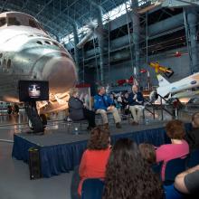 Three Museum staff members host a presentation in front of the Space Shuttle Discovery, a white aircraft designed for space travel, during an open house event.