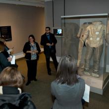 A white female museum conservator speaks to visitors about two tan brown-colored spacesuits on display inside an exhibit about spaceflight.