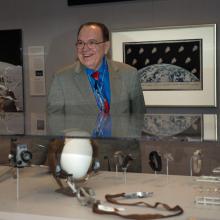 A former NASA manager for astronaut hardware and shuttle crew accomodations smiles behind a display of small spaceflight related objects.