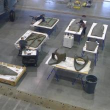 Multiple pieces of a gray, metal aircraft are placed separately on eight tables. Three Museum conservators are performing conservation treatments on separate pieces of the aircraft.