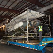 A gray and white metal aircraft, attached to a steel support structure, sits on the long and large bed of a transportation truck. A white tarp protects the top of the aircraft.