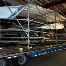 Diagonal back view of the Horton Ho 229 V3, a gray and white metal aircraft, loaded onto a truck for transport. The top of the aircraft is covered by a large white tarp for protection,