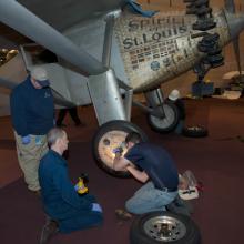 Three Museum staff members work to change tires on the Spirit of St. Louis, a silver monoplane.