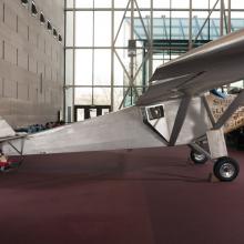 Side view of the Spirit of St. Louis, a silver-colored monoplane, on the floor of the Museum. Registration number "N-X-211" is painted on the tail above the make of the plane, "Ryan MVP".