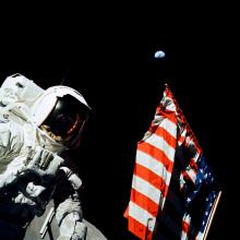 Astronaut Harrison Schmitt stands next to the U.S. Flag planted on the Moon. The top of the red, white, and blue flag is pointed towards Earth.