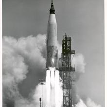 A Mercury-Atlas rocket launching from a launch pad.