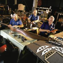 Three restoration volunteers are seated and smiling while working on aircraft parts. A volunteer in the foreground holds sandpaper over the center of a large wood propellor. 