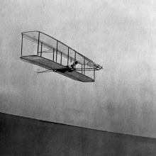 A biplane gliding in the air down a hill. A person lays on top of the bottom wing, perpendicular to it, as they pilot the craft.