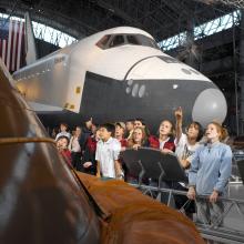 Students stand and point at artifacts hanging above with the Space Shuttle Enterprise in the background.