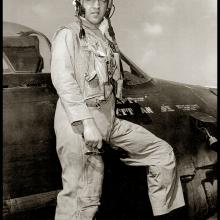 Bill Daniels, a man, stands on the wing of a Grumman F8F Bearcat while wearing aviator gear.