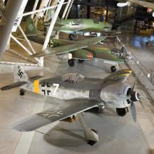 Three German, military aircraft from World War II are displayed next to each other at the Udvar-Hazy Center. The closest aircraft is a silver-colored aircraft and the two aircraft behind it are olive drab green.