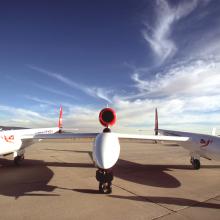 Front view of wide, low winged white aircraft sitting on tarmac with blue sky in the background.