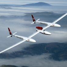 View from above of wide white, low-winged aircraft in flight over cloud-topped mountains.