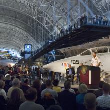 A former director of the National Air and Space Museum speaks in front of a crowd as he welcomes a white fighter jet located behind him at the Udvar-Hazy Center.