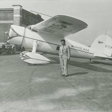 Wiley Post stands on the side of his Winnie Mae aircraft, under its left wing. 