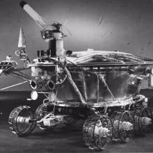 Black and white image of a robot lunar rover with a pie-shaped base and multiple probes, cameras, and antennae attached to the base.