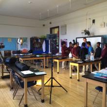 A group of people stand together in a classroom setting on the right while a person films on the left.