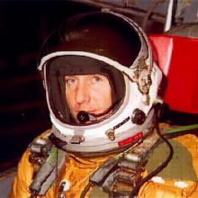 Col. Richard Graham, a white man, sits in an orange and green flight suit and white helmet.