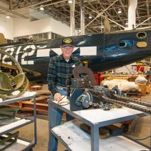 Charles French, a white male veteran, poses in the Restoration Hangar of the museum in front of the Helldiver.