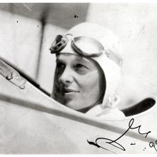 Amelia Earhart sits inside the cockpit of an aircraft. She is wearing an aviator hat and goggles.