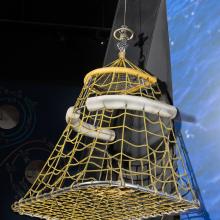 Quadrilateral-shaped metal frame with yellow fabric cords and two white pontoons near top hanging in musuem