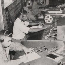 Men sit with headphones on and devices at the center of a table. 