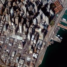 Close-up, satellite view of San Francisco, with high rise buildings to the north and smaller buildings and the San Francisco-Oakland Bay Bridge to the south.