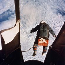 A man jumps out of a gondola of a balloon. You can see him in free fall and the cloud cover below.