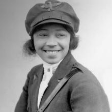 Bessie Coleman, a woman who appears to be in her late 20s, smiles at something to the right of the camera. She is wearing a military-like uniform, with an emblem that reads "B.C." on her cap.