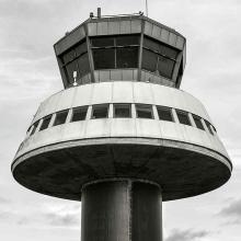 Partial view of an air traffic control tower at Barcelona El-Prat Airport. This tower features a dark-colored cylinder base with a conical-shaped first floor. A second floor features at least four different perspectives for observation.