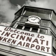 Front view of an air traffic control tower at Cincinnati Municipal Airport. The base is mostly not visible but a sign stating "Welcome to Cincinnati Lunken Airport" is placed under the windows. Above the windows, a clock is visible.