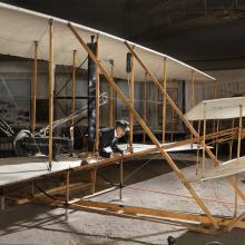 Front of 1903 Wright Flyer with model of Wright Brother laying flat in aricraft