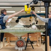 Two Smithsonian employees position an X-ray scanner on top of a large, dish-shaped spaceship studio model.