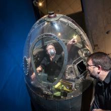 Two white male museum specialists work together to remove a partial fuselage of a historic World War II bomber aircraft from public display in the Museum.