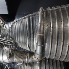 Side view of a rocket engine on display in the museum. One large, conic-shaped nozzle is attached to a fuel manifold pipe at the center of the nozzle. Cooling tubes are attached throughout the nozzle.