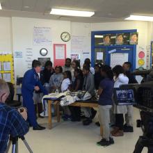 An astronaut wearing a blue full-body suit speaks to several students inside a classroom. Multiple camerapeople are filming the interaction.