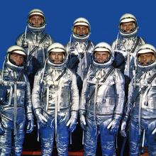 These seven individuals composed the first group of astronauts announced by the National Aeronautics and Space Administration. They stand in shiny metallic suits and smile at the camera.