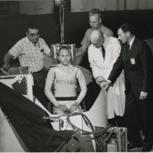 Man sits inside a centrifuge pod, four others surround him. 