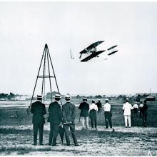 Black and white photo of the Wright Flyer in flight with crowd of onlookers below. 