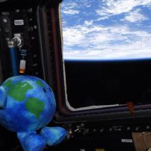 Celestial Buddies plush Earth toy is floating in the international space station. It is shaped and colored like a small Earth with a suprised expression. It is looking out a window towards Earth. 