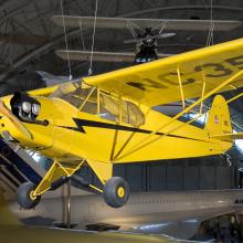 A Piper J-3, a yellow monoplane with one engine, is displayed from a suspended position inside the Museum.