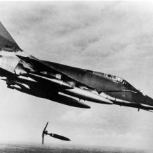 A black and white photo of a North American F-100D aircraft in flight dropping a "Snake eye" bomb on a suspected Viet Cong position in 1966. 