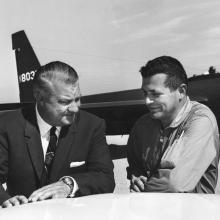 Two men, one in a suit, one in a flight suit, stand in front of an airplane.