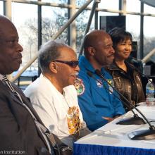 three men and one woman sitting at table with microphones