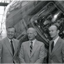 Black and white of Armstrong, Aldrin, and Collins stand beside the Apollo 11 command module.