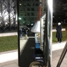 Image of a rear view mirror. In the mirror you can see the outline of the command module on a truck flat bed. 